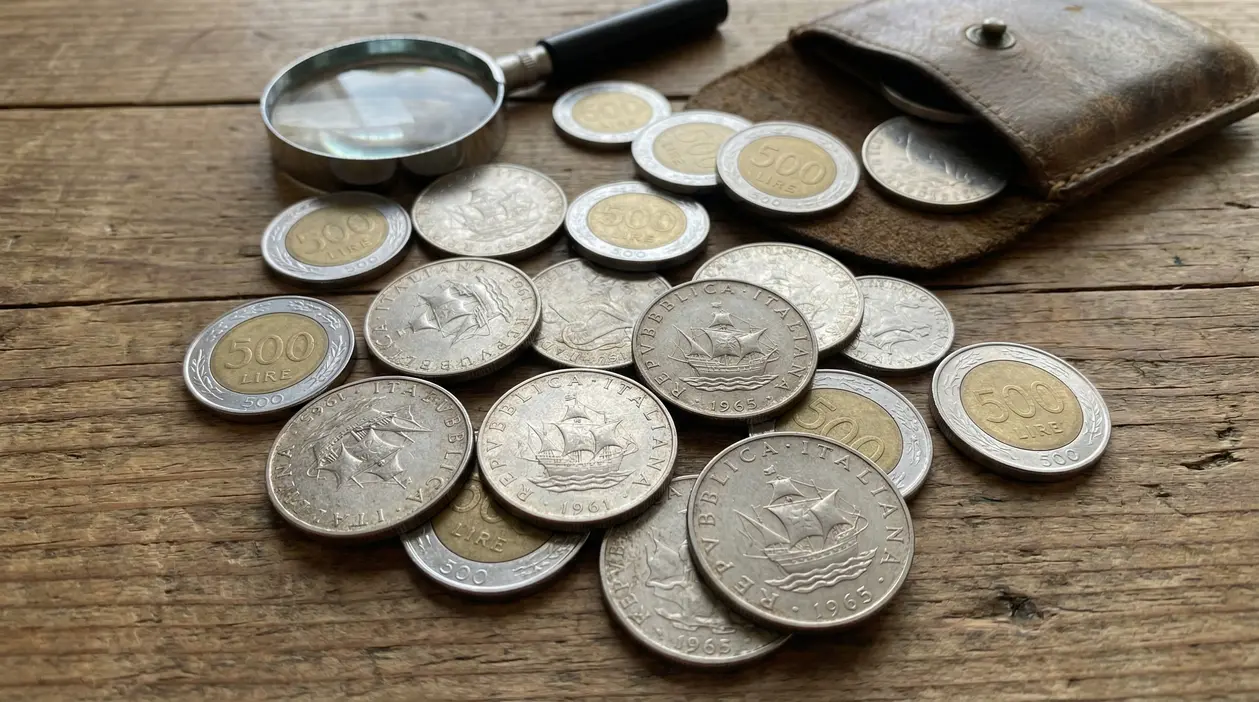 Monete da 500 lire italiane sparse su un tavolo in legno con lente d'ingrandimento e portamonete